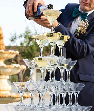 Groom filling a pyramid of glasses with champagne at outdoor garden in wedding ceremony