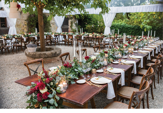 in backyard of villa in Tuscany there is a reception wooden table decorated with cotton and eucalyptus compositions, glasses, candles and plates are placed on table