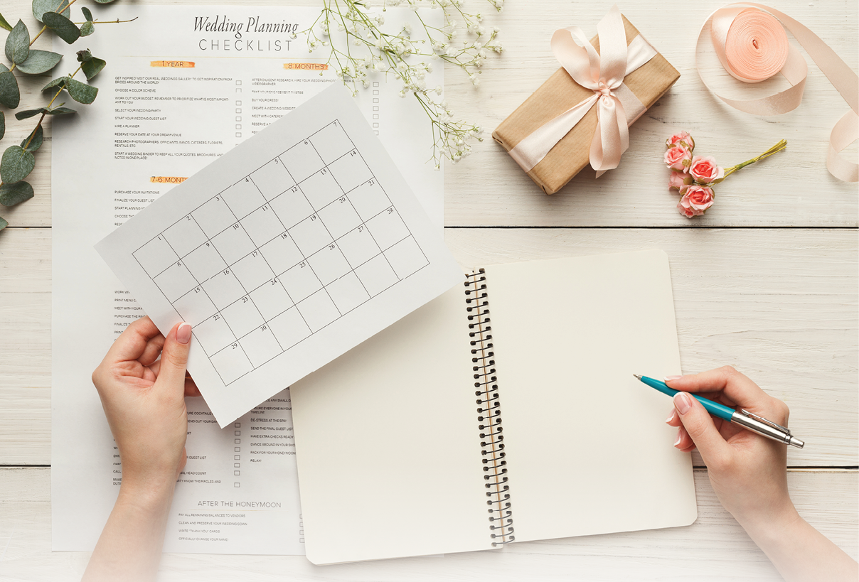 Wedding background with checklist and calendar. Female hands arranging marriage, filling in planners on white wooden table with lots of tender bridal stuff, top view