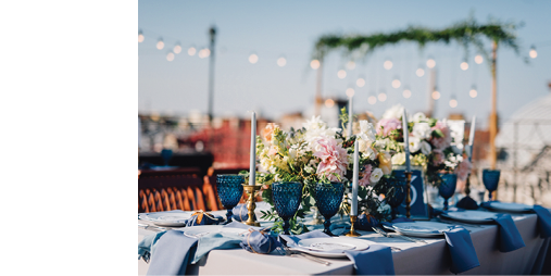 Long dinner tables covered with grey and blue cloth, served with porcelain and blue glasses and rich decorated with flowers stand on the roof of a house  Bokeh lights on baclground