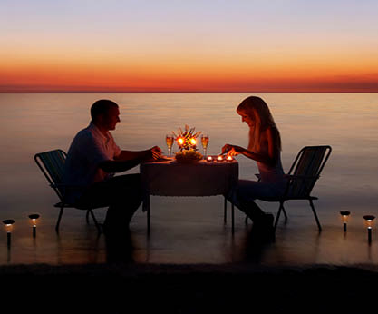 Loving couple share a romantic dinner with candles and lanterns light way at sea beach in water against wonderful sunset